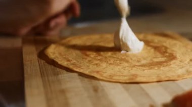İnce, renkli krepler. Philadelphia krem peynirini, tahta bir tahtanın üzerindeki sarı krepi doldurur gibi dağıtan kadınların yakın plan fotoğrafları. 4K video.