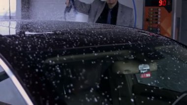 Otomobil yıkamada yüksek basınçlı su teçhizatıyla araba temizleme işlemi. Vitiligo giysili genç adam arabasını foarm ile temizliyor. 4K video