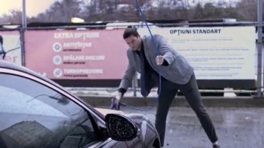 Otomobil yıkamada yüksek basınçlı su teçhizatıyla araba temizleme işlemi. Vitiligo giysili genç adam arabasını foarm ile temizliyor. 4K video