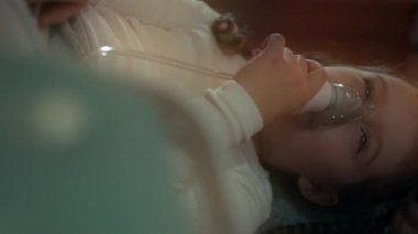 Kanepede otururken kompresör astım spreyi kullanarak nefes alan güzel bir kızın dikey yakın plan portresi. Beş yıl oldu. Yüzünde nebulizör maskesiyle salin buharı soluyan bir kız. Anne!