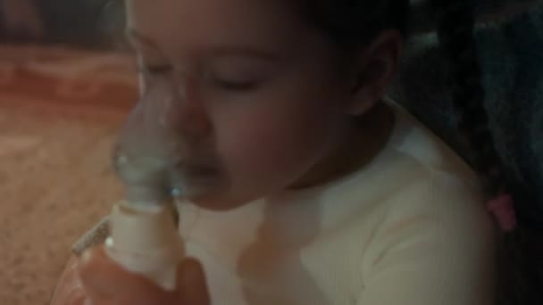 ソファに座っている間、女の子はコンプレッサー吸入器を使用して吸入を行います。5歳。顔にネブライザーマスクで生理食塩水の蒸気を吸い込む少女。母は娘が家で吸入するのを助ける — ストック動画