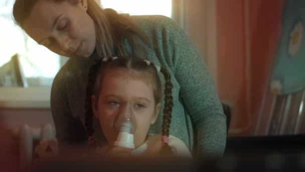 Piękna dziewczynka sprawia, że inhalacja za pomocą inhalatora sprężarki podczas siedzenia na kanapie. 5 lat. Dziewczyna wdychająca sól fizjologiczną z maską nebulizatora na twarzy. Matka pomaga córce w inhalacji — Wideo stockowe