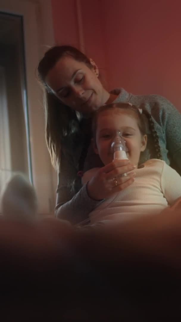 Verticaal közelkép portré gyönyörű kislány, hogy inhalációs kompresszor inhalátor, miközben ül a kanapén. 5 év. Egy lány sóoldatot lélegez be porlasztó maszkkal az arcán. Anyám! — Stock videók