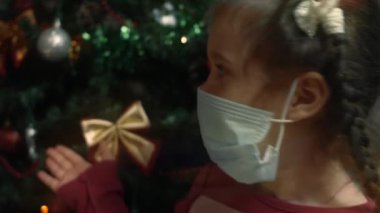 Mutlu küçük kız Noel ağacının arka planında bir Noel şarkısı söylüyor. Beş yıl oldu. COVID-19 salgını sırasında koruyucu maske takan bir kız. 4K video