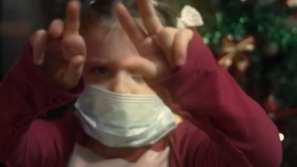 Пять лет. Девушка показывает сердитые эмоции на фоне Рождественской ёлки, недовольная тем, что ей приходится носить защитную маску. 4K видео — стоковое видео