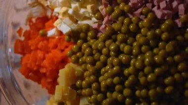 Geleneksel Rus salatası Olivier 'i hazırlıyorum. Cam bir kasenin içindekiler - yeşil bezelye, patates, sosis, havuç, yumurta. 4K video. Üst görünüm