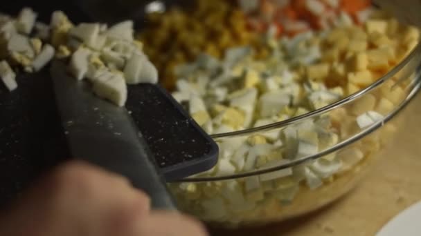Adicionando fatias de ovos cozidos em uma tigela com salada. Preparação de salada de palitos de caranguejo, queijo, pepino, milho enlatado e ovos na cozinha em casa — Vídeo de Stock