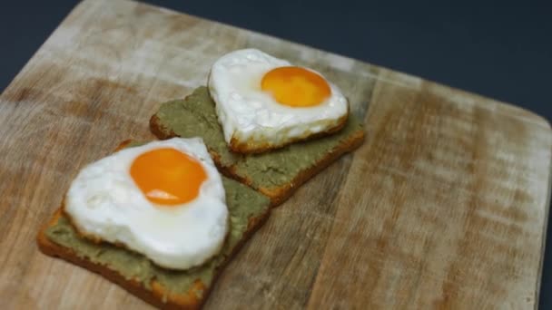 木の板にアボカドを砕いたトーストにハート型の卵。3月の驚きの朝食を調理します。芸術的撮影 — ストック動画