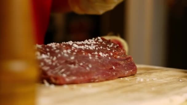Şef çiğ bifteğe deniz tuzu sürüyor. Mutfak masasında et hazırlayan fırıncı çeşitli sebzelerle birlikte 4K görüntüler hazırlıyor. — Stok video