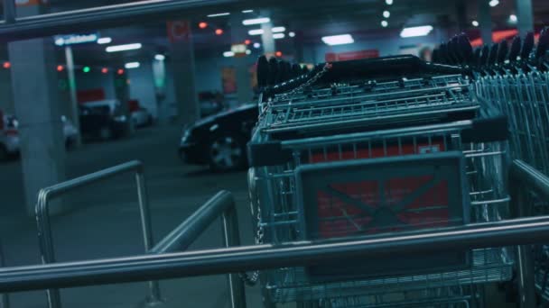 Metalen lege winkelwagen links op overdekte ondergrondse parking van winkelcentrum. Supermarkttrolley met wazig geparkeerde auto 's op de achtergrond. 4k nacht video verkeersbord — Stockvideo