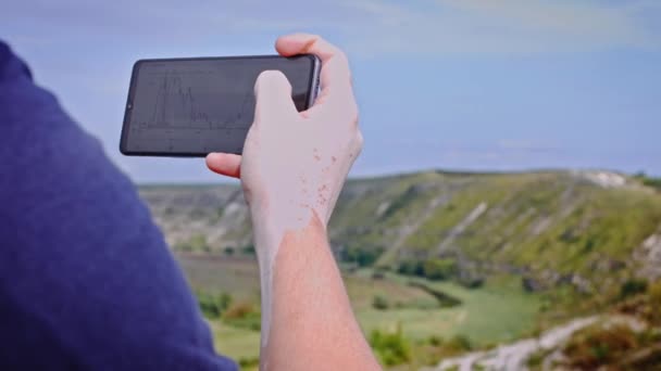 男は電話でBitcoin交換を見ています。岩の上の場所にあります。信仰の象徴。4kビデオ — ストック動画