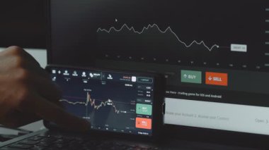 Arkaplanda bir grafiği gösteren bir bilgisayar monitörüyle ticaret uygulaması arayüzü gösteren akıllı telefon.