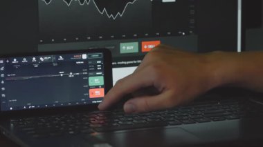 Tüccar, arka plandaki bir dizüstü bilgisayarla çevrimiçi ticaret platformu arayüzü ile etkileşime geçiyor.