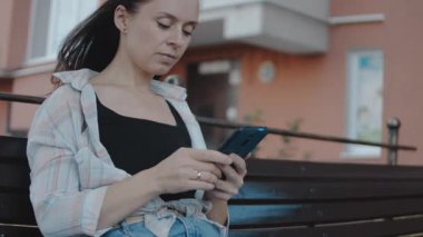 Genç bir kadın bankta tek başına oturuyor ve elinde bir akıllı telefon tutuyor..