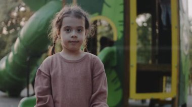 Küçük kız çocuk parkında dikilip kameraya bakıyor..