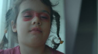 Gülümseyen küçük kız yüzünü ruj ve göz farı ile boyamış ve kameranın önünde soytarılık yapıyor..