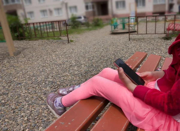 A girl in pink clothes plays on the phone outside sitting on a bench on the playground in sunny weather