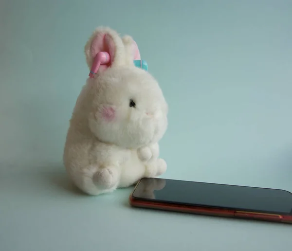 Beyaz bir oyuncak tavşan kulaklıkla mavi arka planda akıllı telefonun yanında oturuyor. — Stok fotoğraf