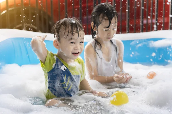 Şirin küçük oğlan ve kız kardeş şişme havuzda baloncuklarla ve renkli toplarla oynarken eğleniyorlar. Şişme ve kabarcık havuzları, yazın yüzme, suda oynayan mutlu çocuklar.
