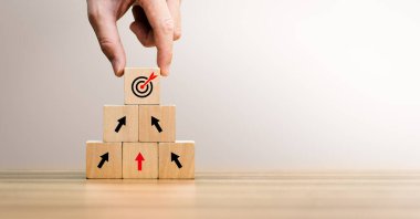 Elle tahta blokları yukarı doğru istifleyerek, okları ve hedef simgeyi gösteren iş büyüme konsepti. Strateji, hedef başarısı, kalkınma, planlama, liderlik ve uzun vadeli başarıyı temsil eder.