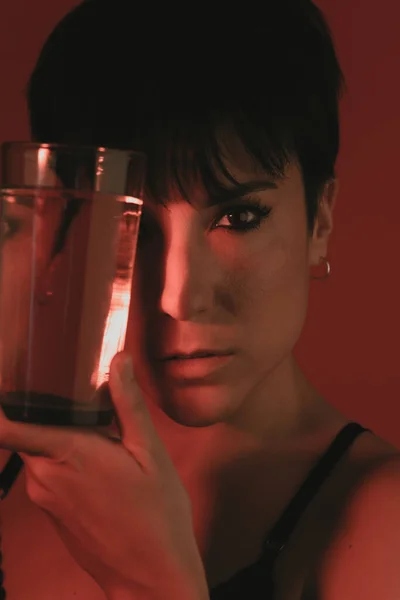 Stüdyoda bir bardak suyla yüzünü örterken kameraya bakan bir kadının portresi.