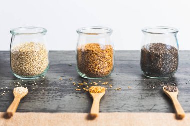 Üç bardak kinoa, keten ve siyah arka planda üç ahşap kaşık bulunan chia. Kırsal kahverengi bir kumaş öne çıkıyor.