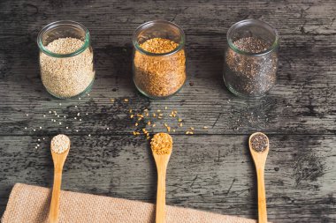 Üç bardak kinoa, keten ve siyah arka planda üç ahşap kaşık bulunan chia. Kırsal kahverengi bir kumaş öne çıkıyor.