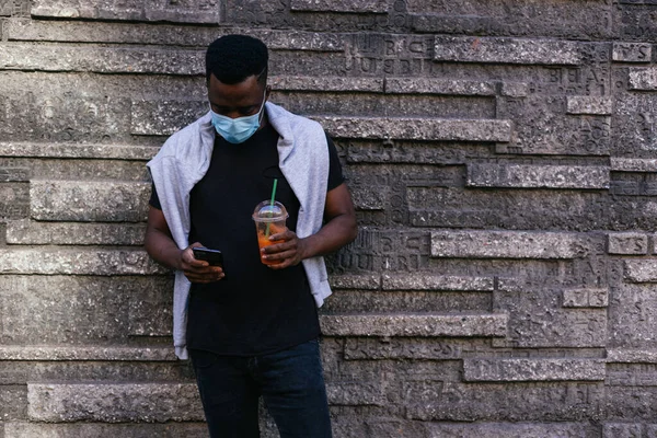 Maskeli Afro-Amerikalı telefonuna bakıyor ve diğer yandan smoothie içiyor.