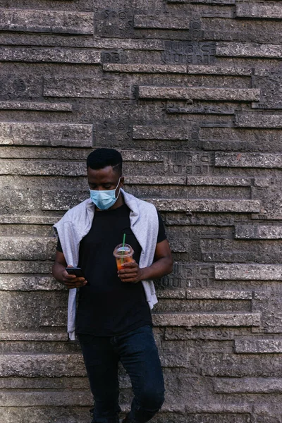 Maskeli Afro-Amerikalı telefonuna bakıyor ve diğer yandan smoothie içiyor.