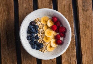 Bir kase meyve, muz, ahududu ve yulaf ve yoğurtlu ahşap arka planda yoğurt.