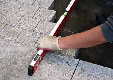 İnşaat eldivenlerindeki bir tuğla ustasının elleri, kentteki bir caddeye kaldırım döşeyen granit kaldırım taşlarının yüzeyini düzleştirmek için bir seviye taşır. Yol yapımı işe yarıyor. Şehrin gelişimi.. 