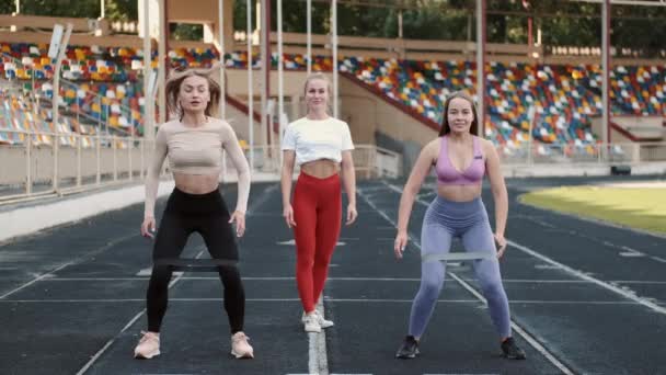 Deportivas y entrenadoras haciendo sentadillas con banda elástica de resistencia en el estadio — Vídeo de stock