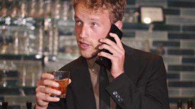 Genç işadamı barda oturmuş viski içiyor ve telefonla konuşuyor.