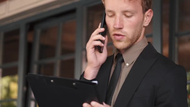 若いビジネスマンは携帯電話で話している間にクリップボードで読む — ストック動画