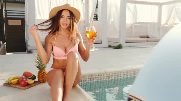 Gorąca opalona kobieta w stroju kąpielowym i słomkowym kapeluszu z koktajlem przy basenie — Wideo stockowe