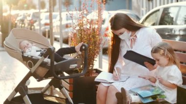 Koruyucu maskeli güzel bir anne ve şehirde bankta oturan iki çocuk.