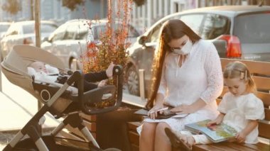 Koruyucu maskeli güzel bir anne ve şehirde bankta oturan iki çocuk.