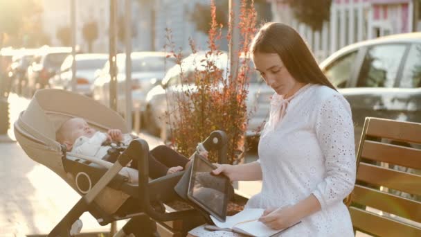 Mor sitter på en pinne med en bebis i barnvagn och läser tidningar — Stockvideo