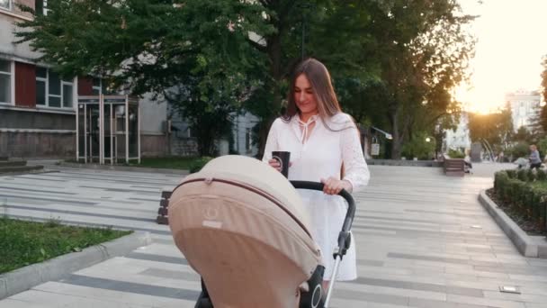 Mooie moeder wandelen met een wandelwagen en koffie op de stoep op zonsondergang — Stockvideo