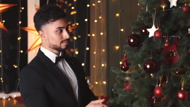 Bonito homem de terno está decorando árvore de Natal com bolas vermelhas — Vídeo de Stock