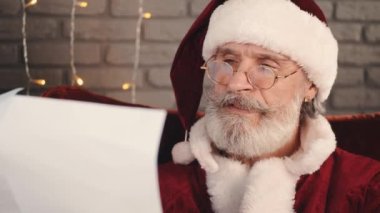 Gözlüklü Noel Baba oturuyor ve mektup okuyor.