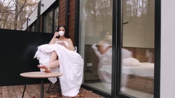 Bella ragazza che riposa e beve caffè seduto sulla terrazza di una loggia in una sedia avvolta in una coperta bianca — Video Stock