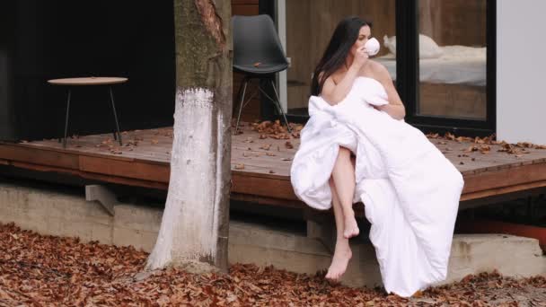 Gadis cantik beristirahat dan minum kopi duduk di teras pondok di kursi dibungkus selimut putih — Stok Video