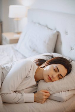 Kadın yatakta, beyaz yastık da odada uyur.