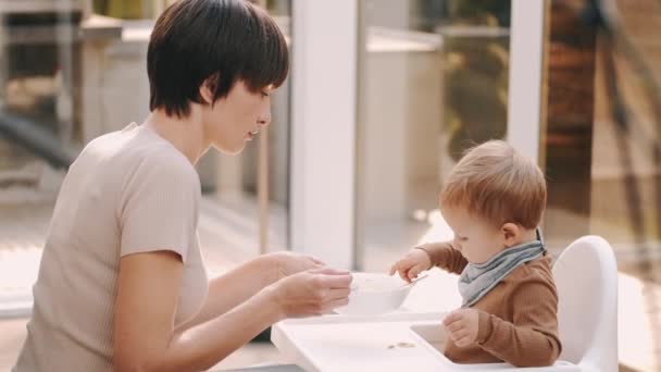 Το παιδί κάθεται σε μια καρέκλα μωρού και τρώει τα τρόφιμα με τη μητέρα — Αρχείο Βίντεο