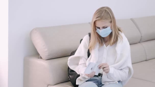 Chica joven con máscara protectora sentada en el sofá en la clínica médica — Vídeo de stock