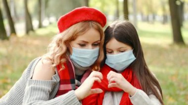 Maskeli anne ve kızı, sonbahar parkında elleri ve kalbi gösteriyor.