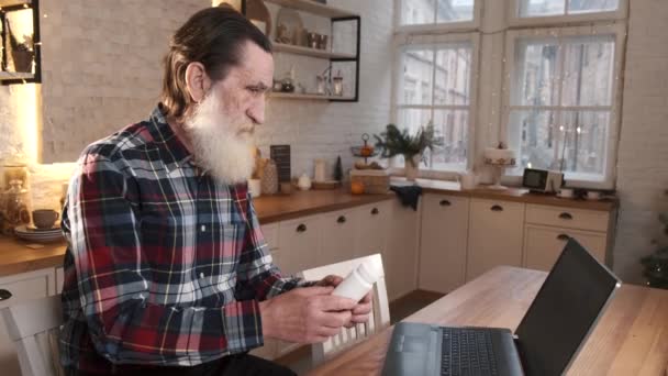 L'homme âgé communique avec le médecin en ligne via la communication vidéo à l'aide d'un ordinateur portable assis à la maison à table pendant la quarantaine — Video