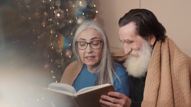 Senioren paar besteden gelukkige tijd thuis het lezen van een boek samen met een versierde kerstboom — Stockvideo