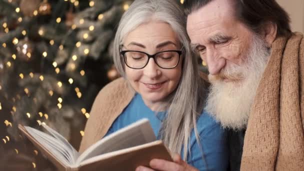 Senioren paar besteden gelukkige tijd thuis het lezen van een boek samen met een versierde kerstboom — Stockvideo